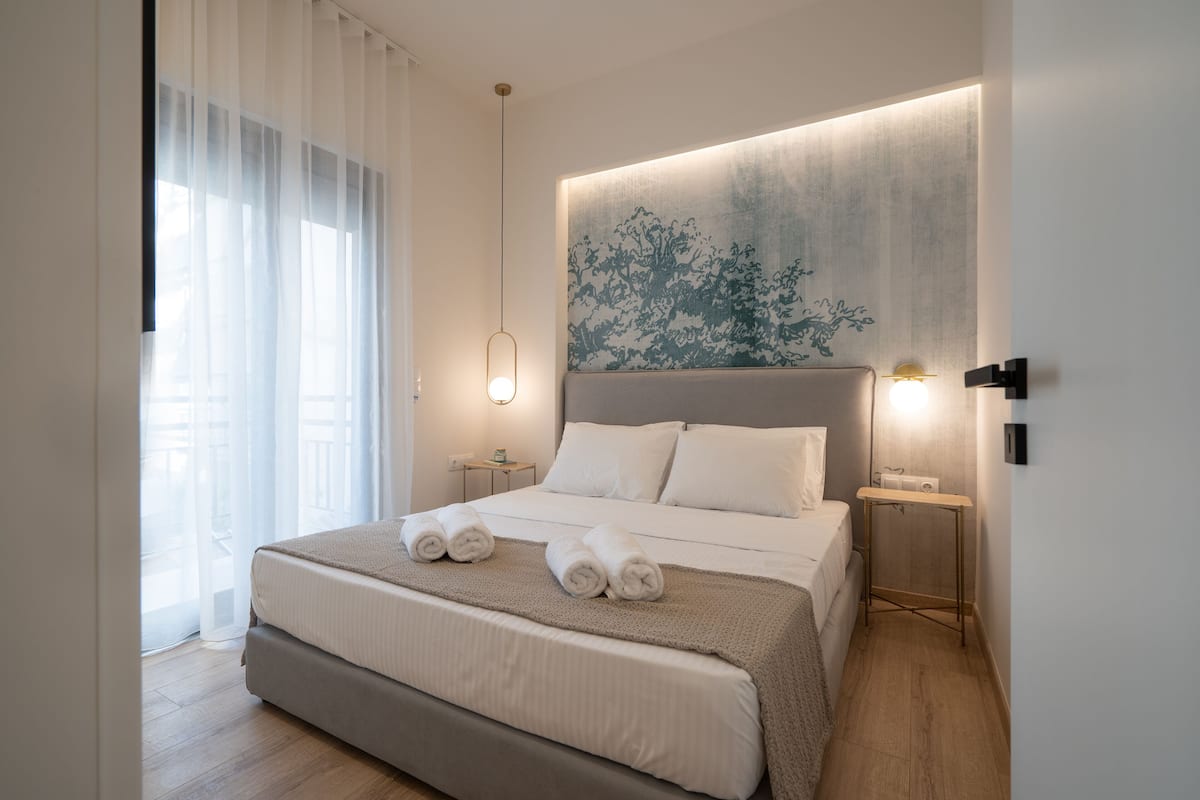 A modern bedroom with a large bed, a bedside table, a wall-mounted light, and a window with sheer curtains.