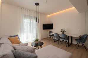 Un salon moderne comprenant un canapé, une table à manger avec des chaises, un téléviseur fixé au mur et une fenêtre ornée de rideaux légers.