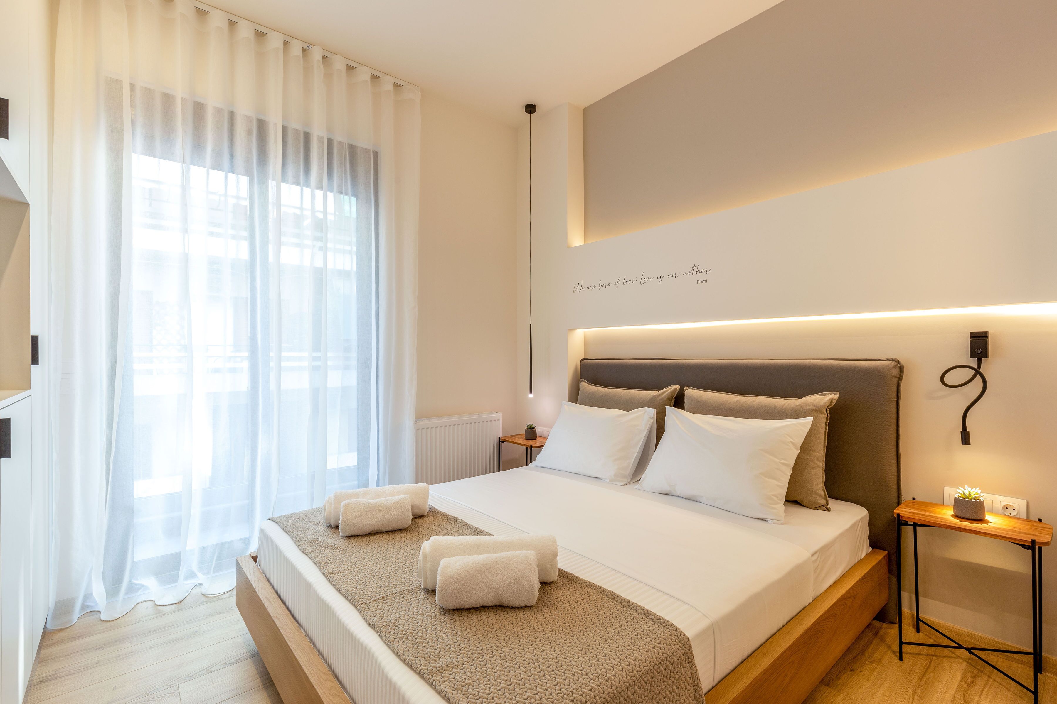 A modern hotel room with a large bed, white linens, a headboard with integrated lighting, a bedside table with a plant, and a view through sheer curtains.