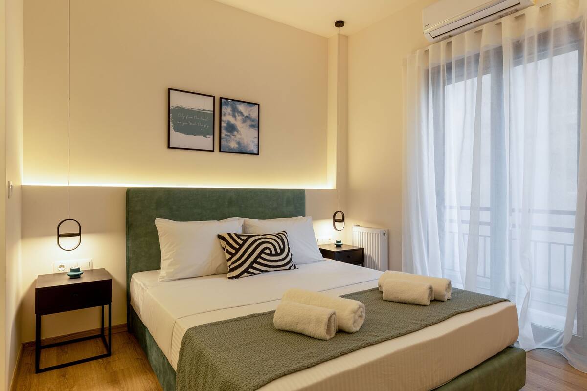 A neatly made bed with a green headboard, white bedding, and two pillows. There are two bedside tables with lamps, a framed picture above the bed, and a window with sheer curtains.
