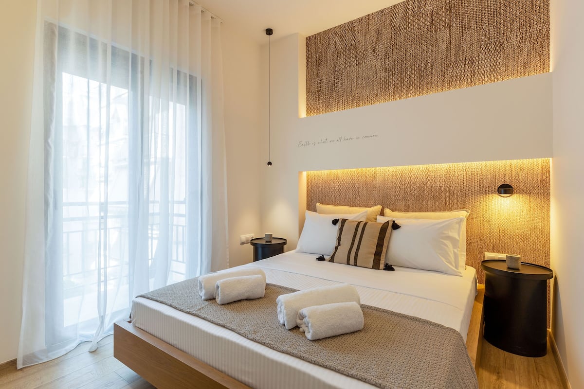 A modern hotel room with a large bed, a bedside table, a wall-mounted light, and a view of the outdoors through sheer curtains.