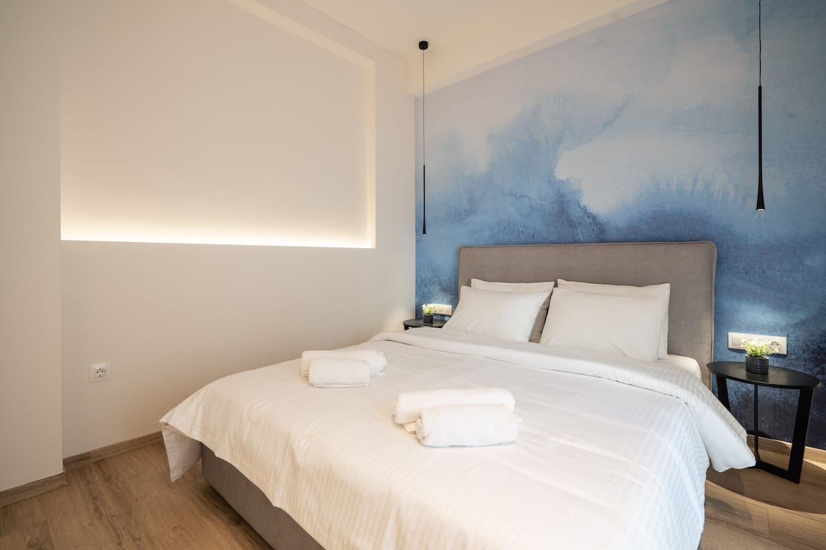 A modern hotel room with a large bed, white linens, a blue abstract wall mural, and a small bedside table with a lamp.