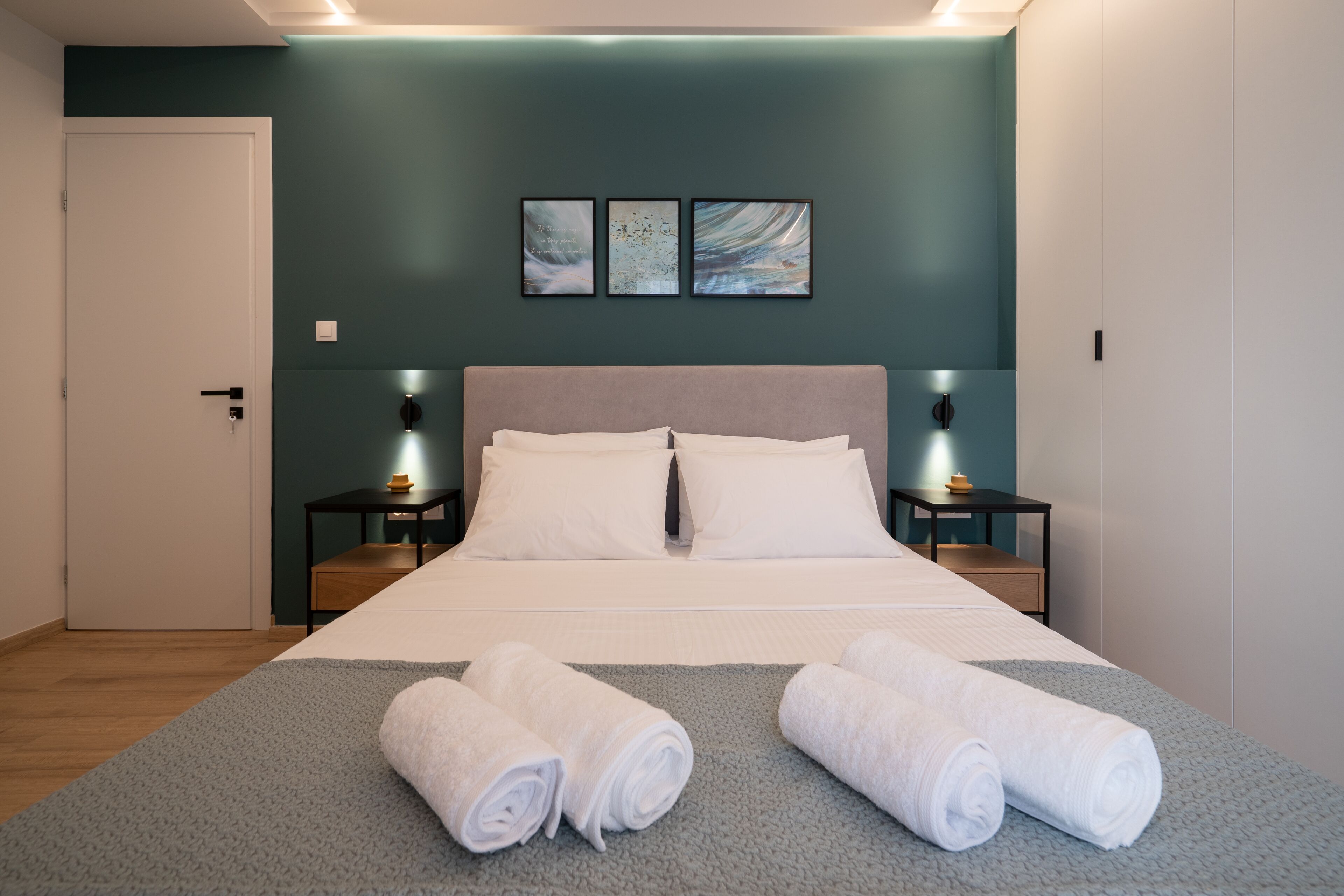 Un lit bien fait, avec du linge de lit blanc et deux serviettes enroulées posées sur un tapis gris, un mur de couleur turquoise orné de trois œuvres d’art encadrées et une porte blanche.