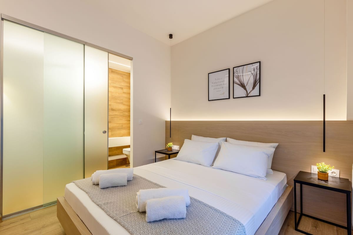 A modern hotel room with a large bed, a wooden headboard, a bedside table with a plant, and a sliding door closet.