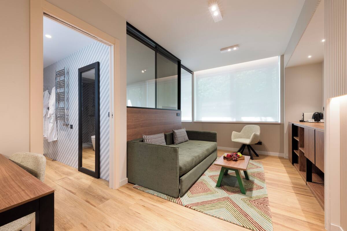 Een moderne, goed verlichte kamer met een bank, een klein tafeltje met fruit, een stoel en een groot raam.