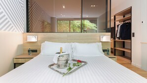 Une chambre d’hôtel dotée d’un grand lit, d’un minibar, d’une table de chevet, d’une petite table, d’une armoire et d’une grande fenêtre donnant sur un paysage verdoyant.