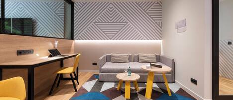 A modern hotel room with a wooden desk, two yellow chairs, a gray sofa with patterned cushions, two small round tables, and a colorful geometric rug.