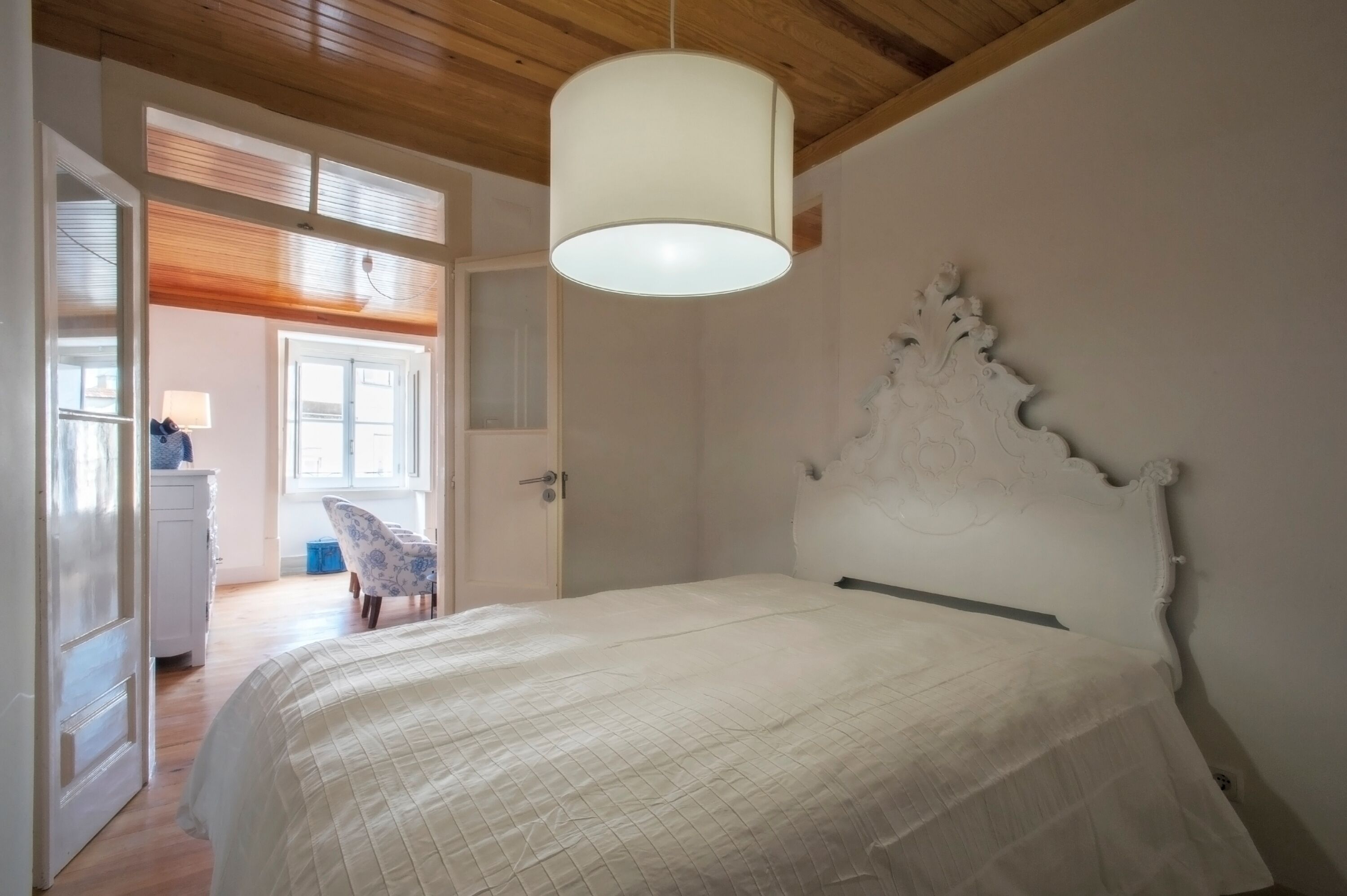 A bedroom with a bed, a white headboard, a hanging light fixture, a chair, and a door leading to another room.