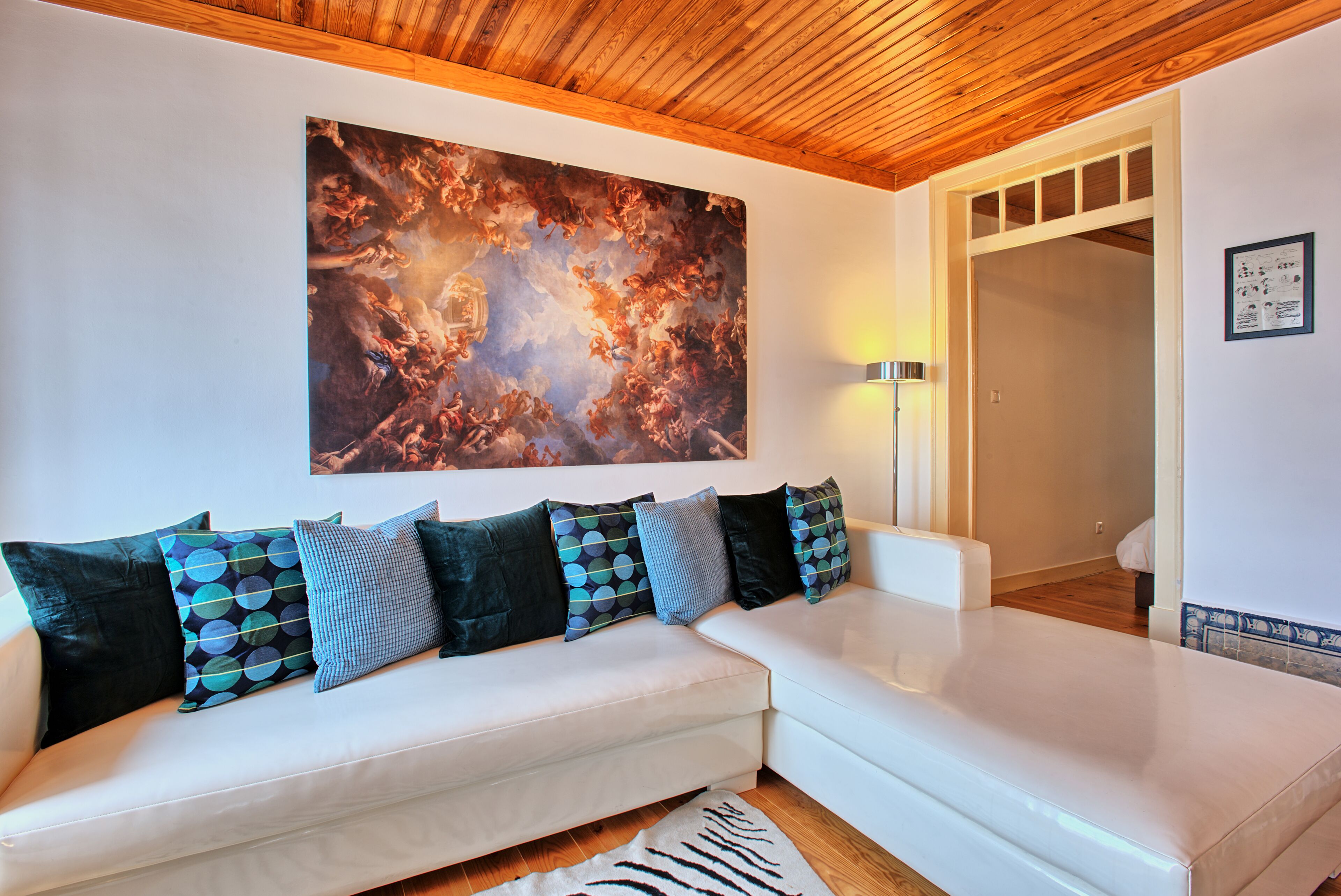 A white sofa with patterned cushions, a large abstract painting on the wall, and a wooden ceiling.