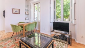 Una sala de estar moderna con una mesa de centro de vidrio, una mesa de comedor de madera con sillas verdes, un televisor de pantalla plana sobre un mueble bajo y un ventanal con vistas a árboles.