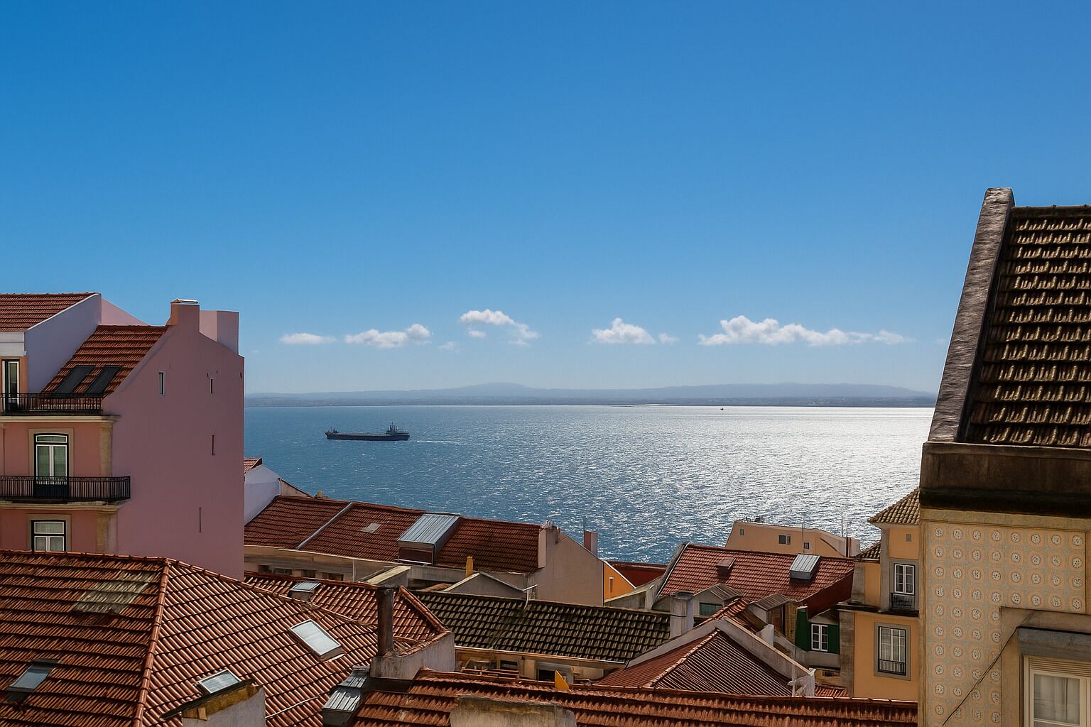 Un paysage urbain côtier avec des bâtiments aux toits de tuiles rouges, un ciel bleu dégagé et un navire sur l’eau.
