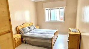 Une chambre à coucher avec un lit, des tables de chevet en bois, une commode et une fenêtre avec des stores.