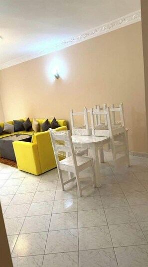 Un coin repas avec une table et des chaises blanches, un canapé jaune agrémenté de coussins et un sol carrelé.