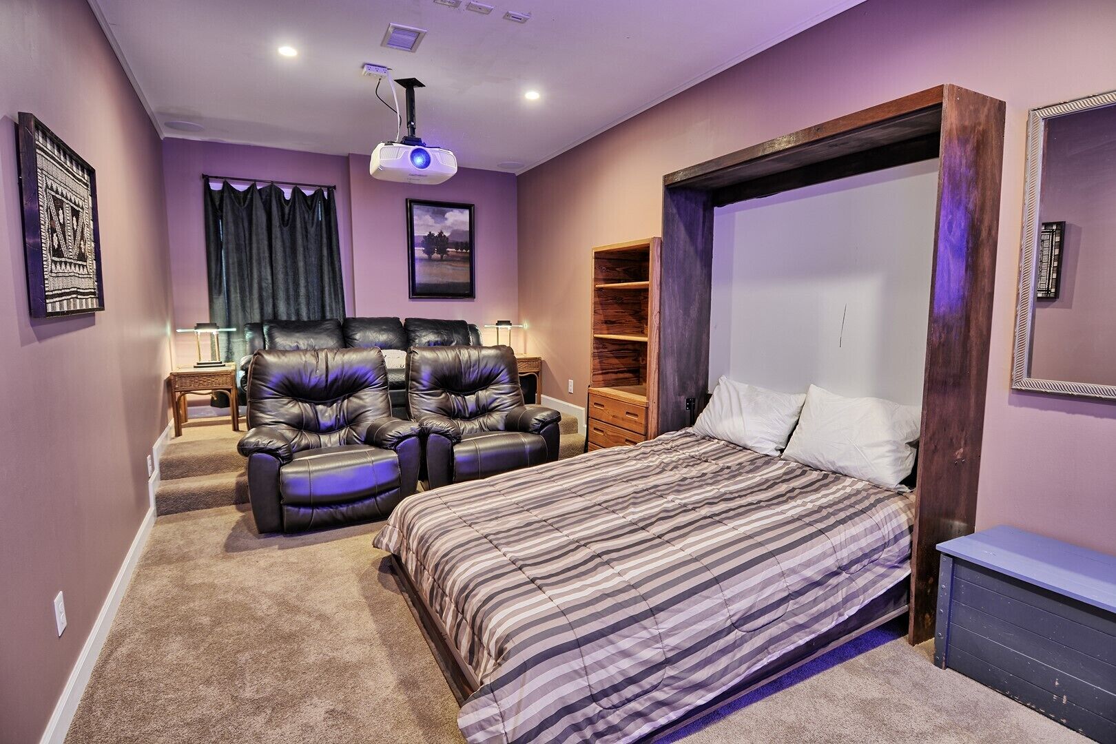 A bedroom with a bed, two leather recliners, a wooden wardrobe, and a ceiling projector.
