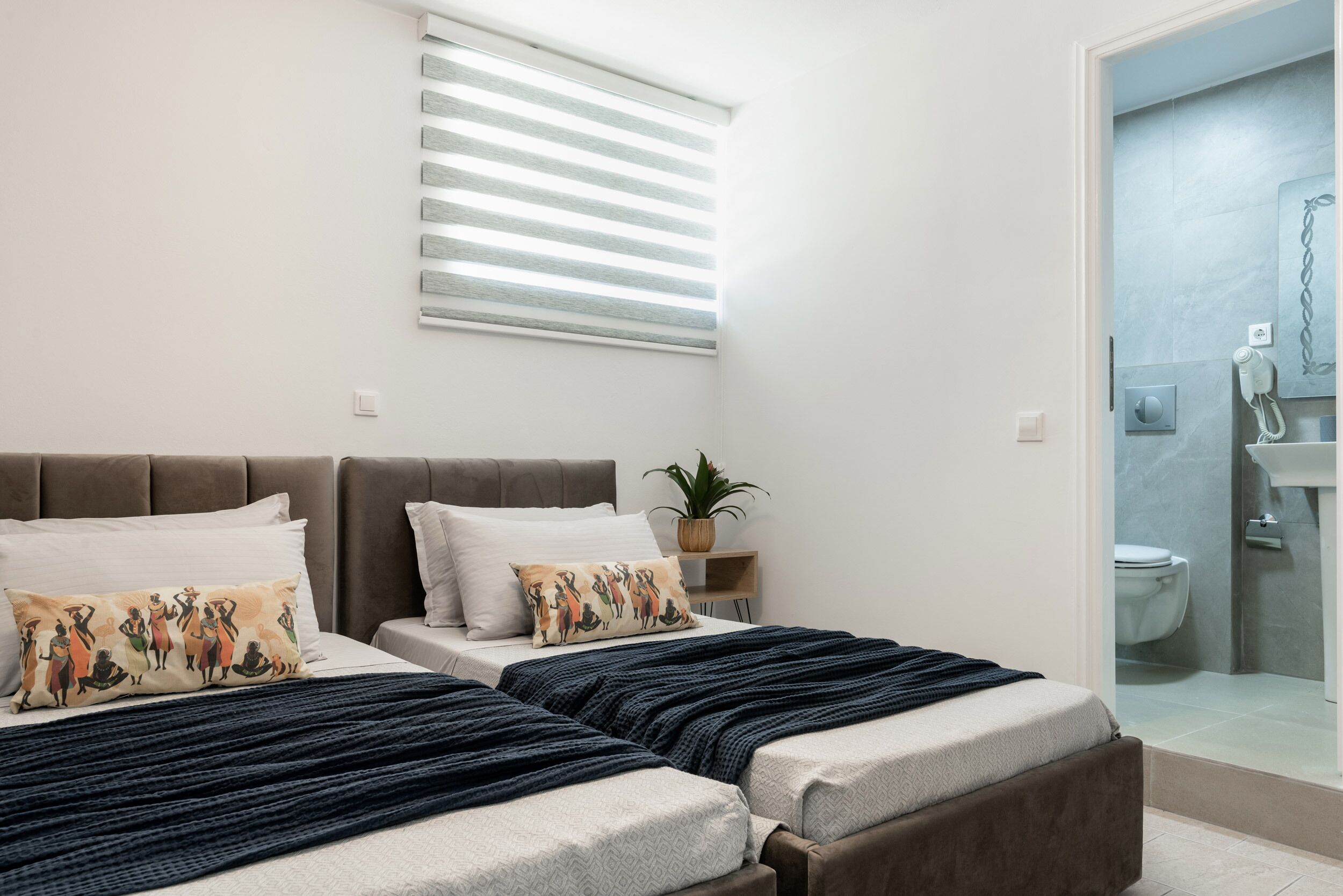 A bedroom with two beds, a bedside table with a plant, and a bathroom visible through a glass door.