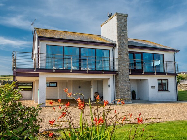 A modern two-story house with large windows, a stone chimney, and a balcony. - The Steading (South Walls)