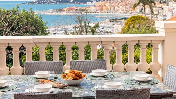 Ein Esstisch mit Tellern, Schüsseln und einer Schüssel Croissants auf einem Balkon mit Blick auf eine Küstenstadt.