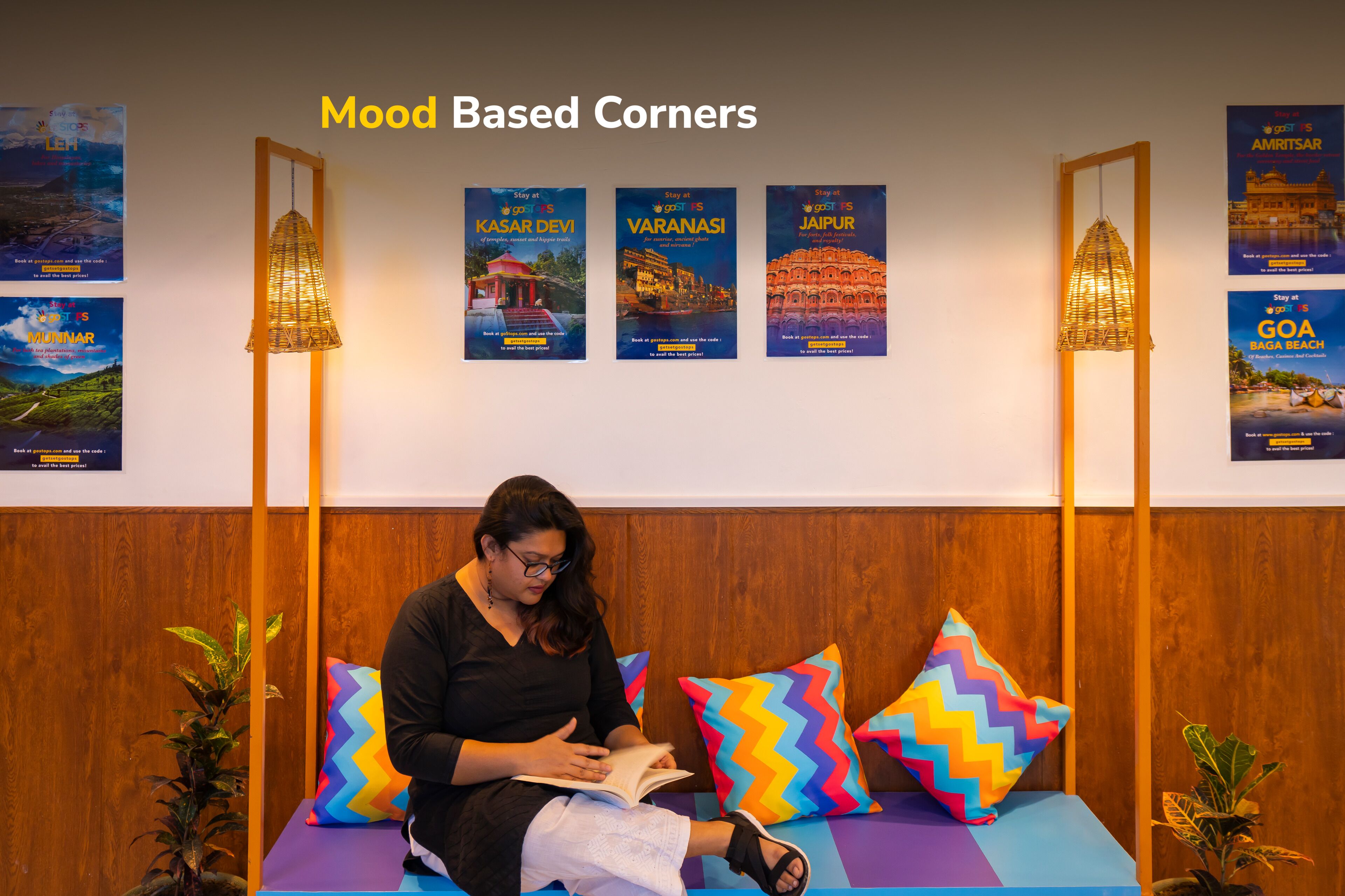 Une personne est assise dans une pièce, lisant un livre. La pièce est décorée avec un mur lambrissé en bois, des coussins colorés à motifs en zigzag et des affiches de voyage accrochées au mur.