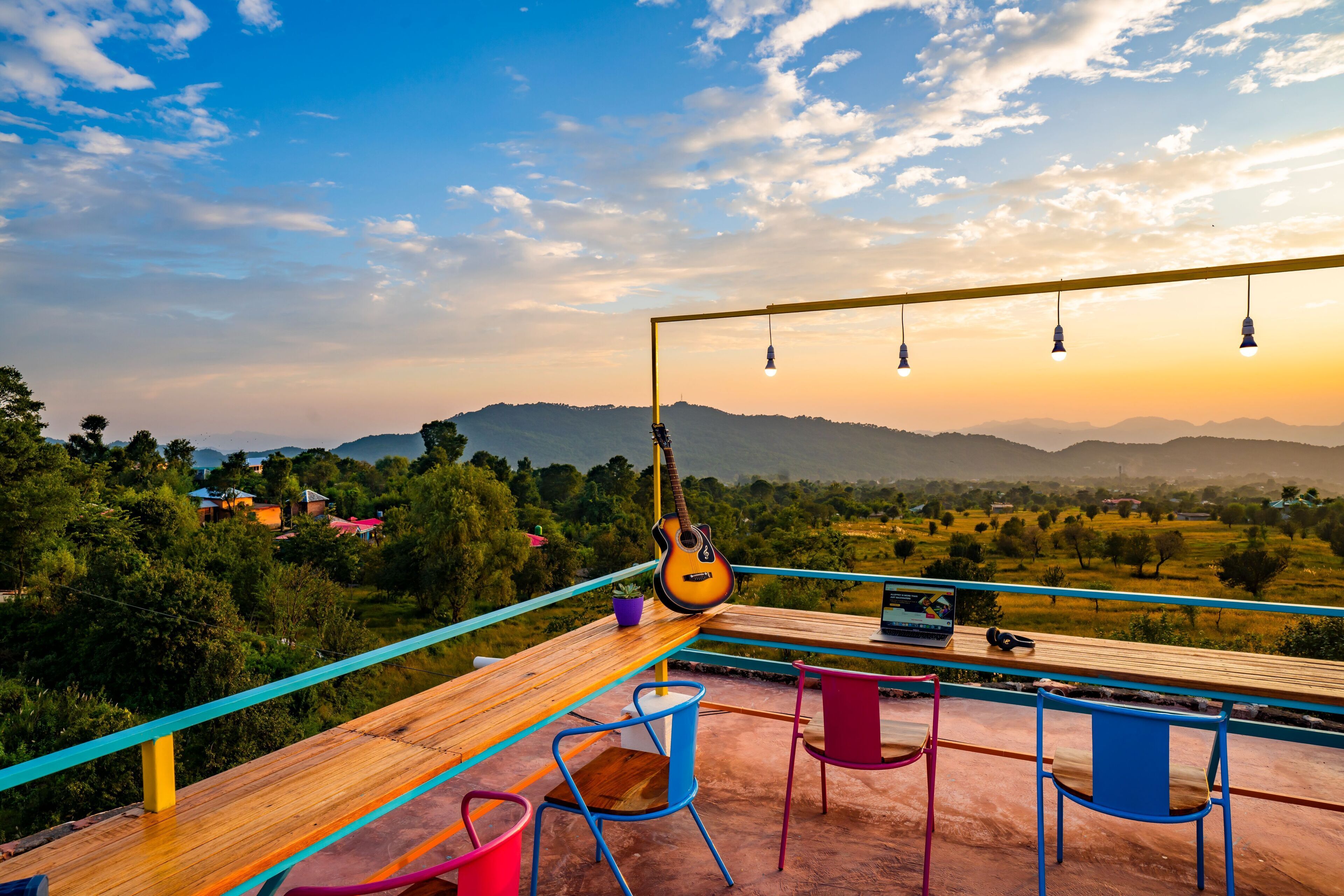 A wooden deck with a guitar, chairs, and a table overlooking a scenic landscape with mountains and trees.