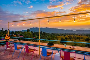 A terrace with a wooden table, colorful chairs, a laptop, a guitar, and hanging lights against a sunset view. - goSTOPS Bir, Landing Site (Baijnath)