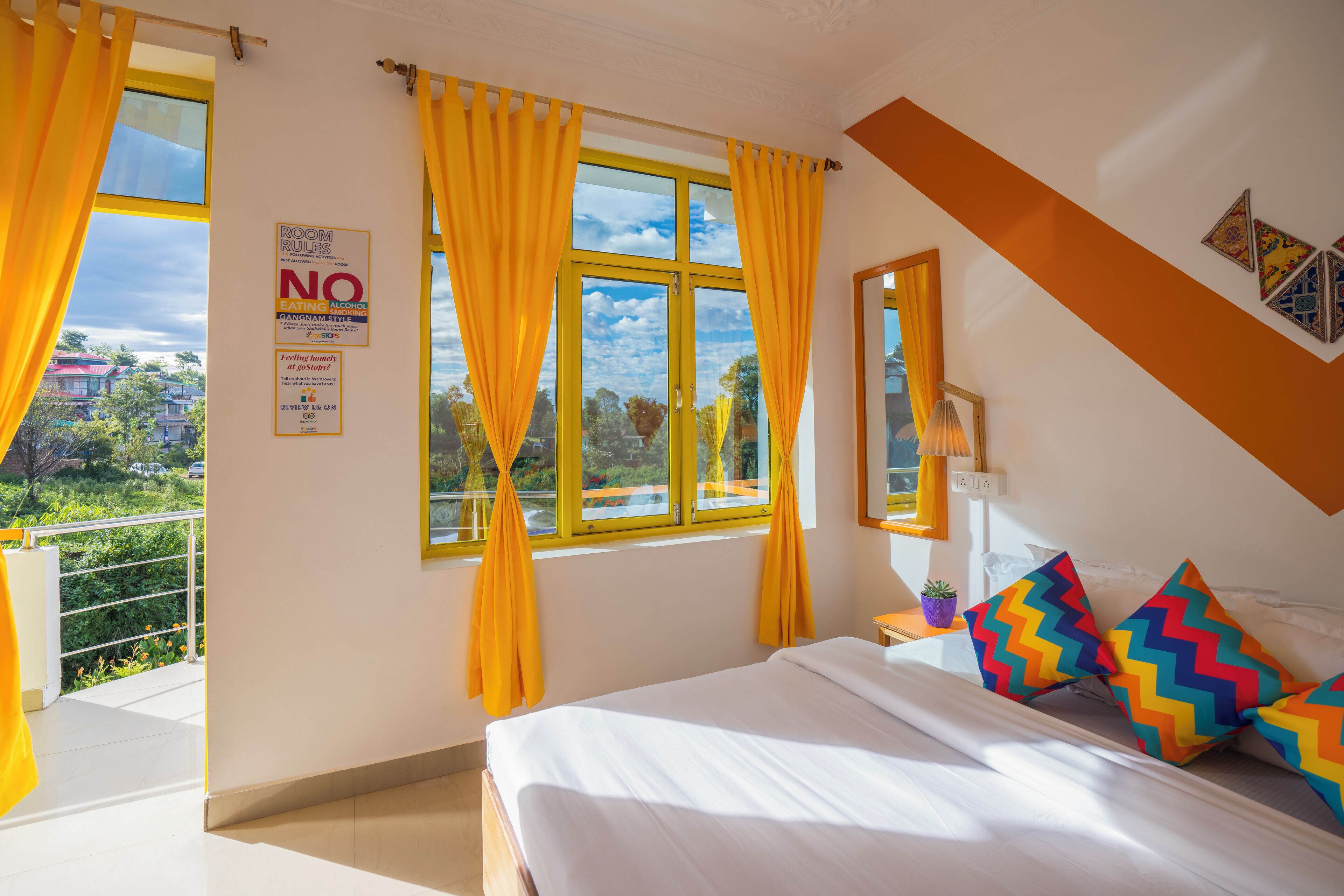 Una habitación de hotel con cama, cortinas amarillas, ventana con vistas, un cartel en la pared y una planta pequeña en la mesita de noche.