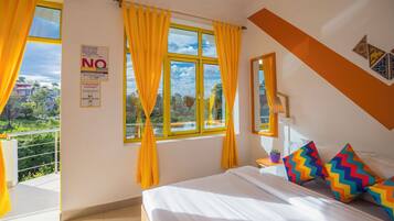 Una habitación de hotel con cama, cortinas amarillas, ventana con vistas, un cartel en la pared y una planta pequeña en la mesita de noche.
