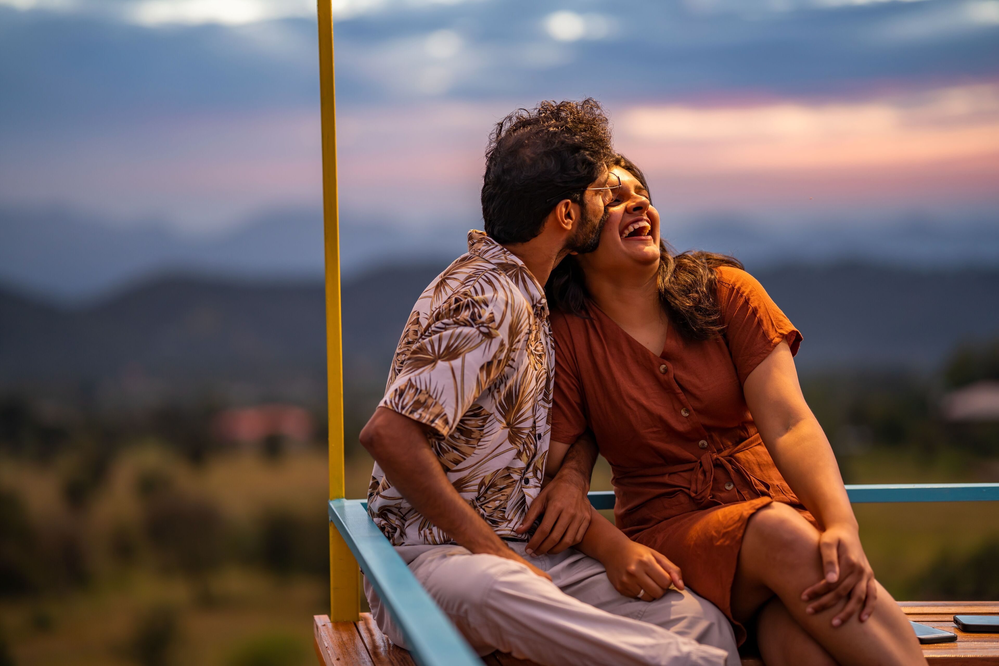 Un couple est assis sur un banc, avec une vue panoramique sur les montagnes et un coucher de soleil en arrière-plan.