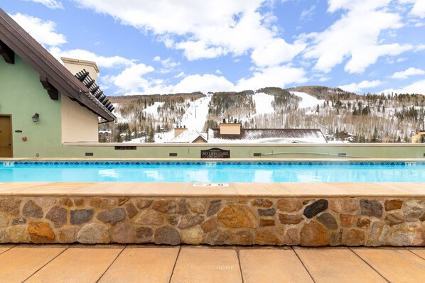 Una piscina con borde de piedra, un edificio con un cartel que dice "MARTIN'S NO LIFEGUARD ON DUTY" y una montaña nevada al fondo.