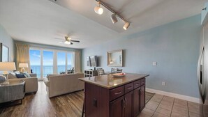 A spacious living area with a large window overlooking the ocean, a ceiling fan, a flat-screen TV, and a kitchen island with cabinets.
