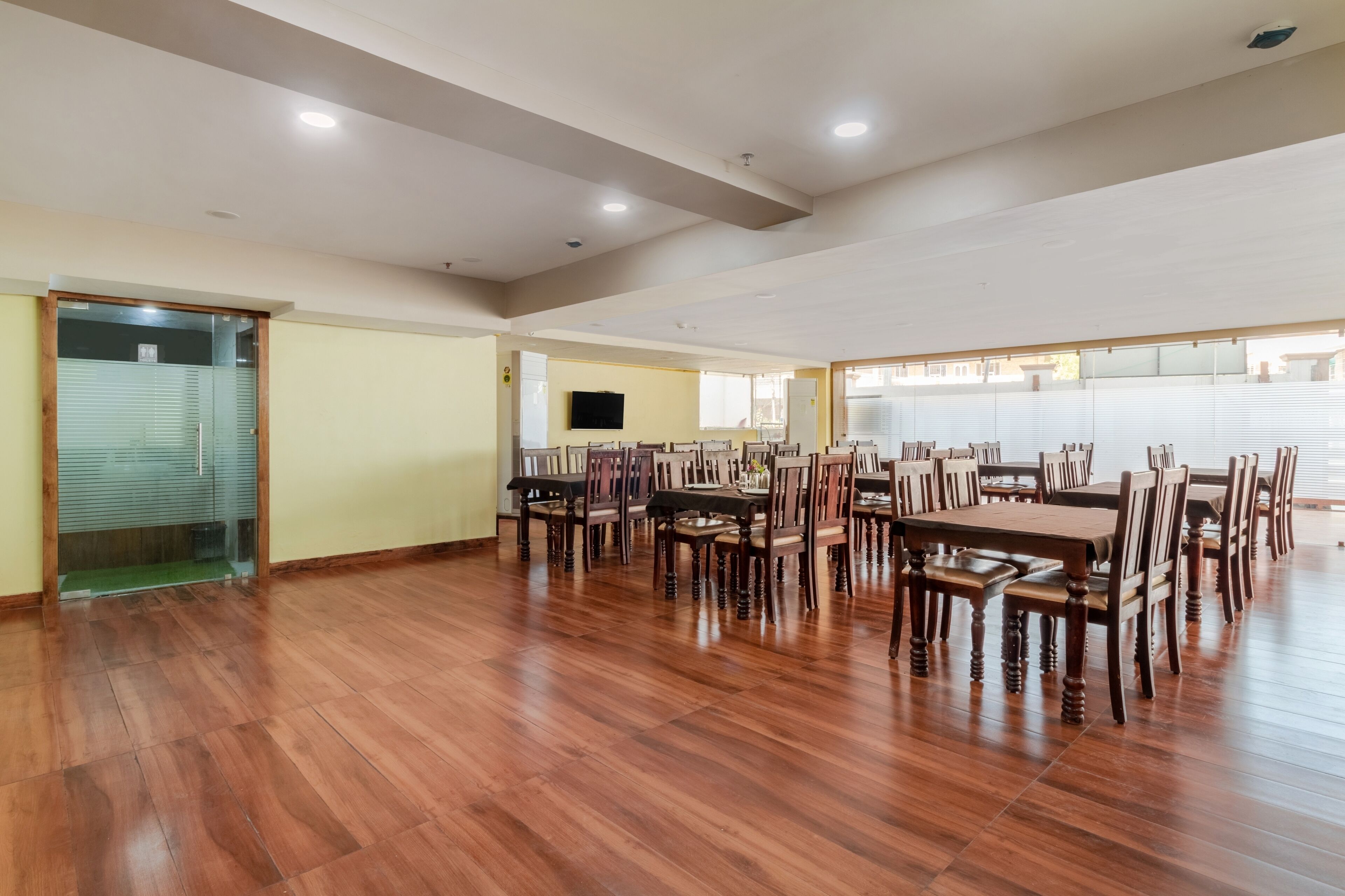 A spacious dining area with wooden tables and chairs, a flat-screen TV mounted on the wall, and large windows with blinds.