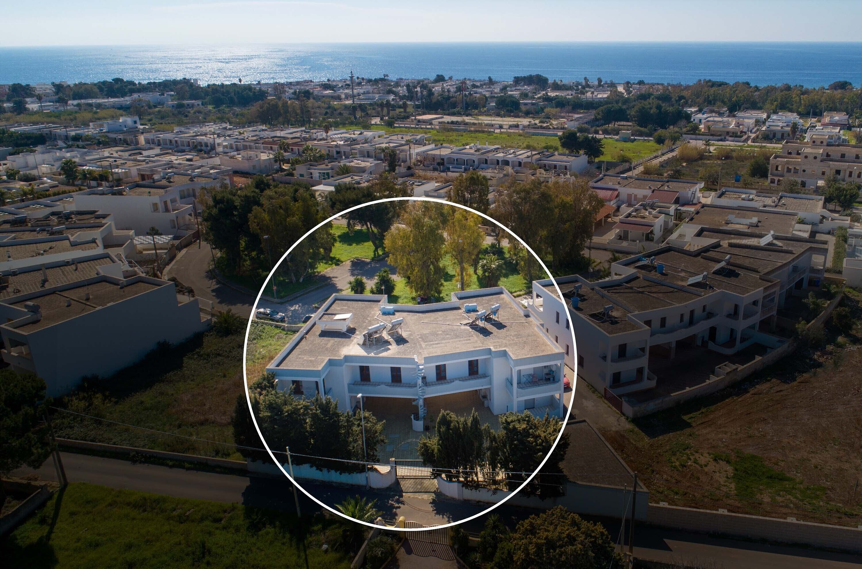 An aerial view of a residential area with a highlighted building featuring a central courtyard, surrounded by other buildings, and a coastline in the background.