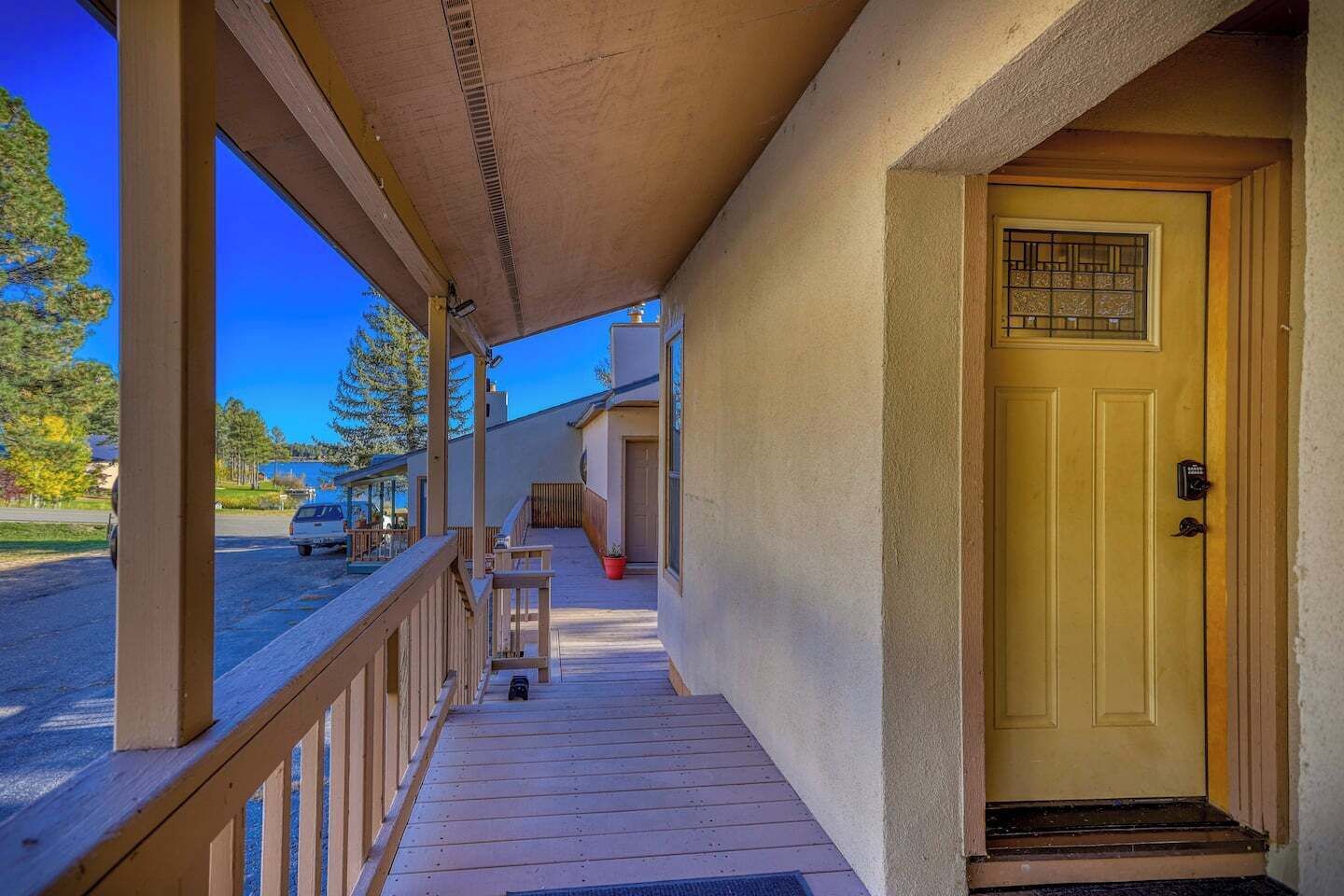 A covered porch with a wooden railing, a yellow door with a window, and a view of a street with a parked car.