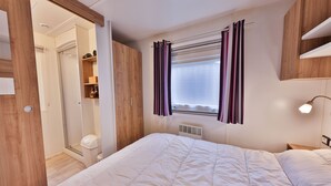 A bedroom with a bed, a window with curtains, a bedside table, and a wardrobe. - Grand Mobil-home 6 persons 3 bedrooms 2 bathrooms camping 3* Parc de la Fecht (Munster (68140))