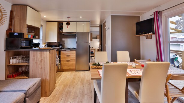A compact kitchen with wooden cabinets, a built-in microwave, and a refrigerator. There is a dining area with a table and chairs, and a TV mounted on the wall. - Grand Mobil-home 6 persons 3 bedrooms 2 bathrooms camping 3* Parc de la Fecht (Munster (68140))