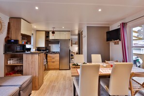 A compact kitchen with wooden cabinets, a built-in microwave, and a refrigerator. There is a dining area with a table and chairs, and a TV mounted on the wall. - Grand Mobil-home 6 persons 3 bedrooms 2 bathrooms camping 3* Parc de la Fecht (Munster (68140))