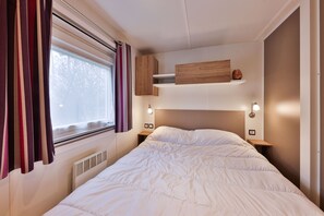 Un lit bien fait, avec du linge de lit blanc, une fenêtre avec des rideaux et une tête de lit dotée d’étagères intégrées.