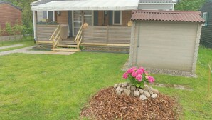 A wooden house with a porch, a small shed, and a flower bed with pink flowers. - Grand Mobil-home 6 persons 3 bedrooms 2 bathrooms camping 3* Parc de la Fecht (Munster (68140))