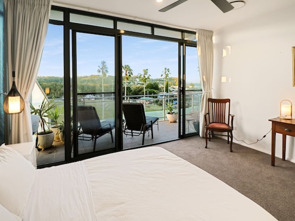 A bedroom with a large bed, a desk, a chair, and a balcony with chairs and a view of a body of water and greenery.