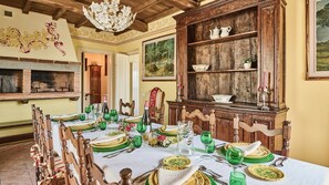 Un comedor con una larga mesa dispuesta para una comida, sillas de madera, un aparador de madera con estantes y una araña.