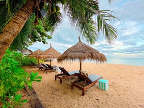 Una playa con sombrillas de paja, sillones de madera y arena.