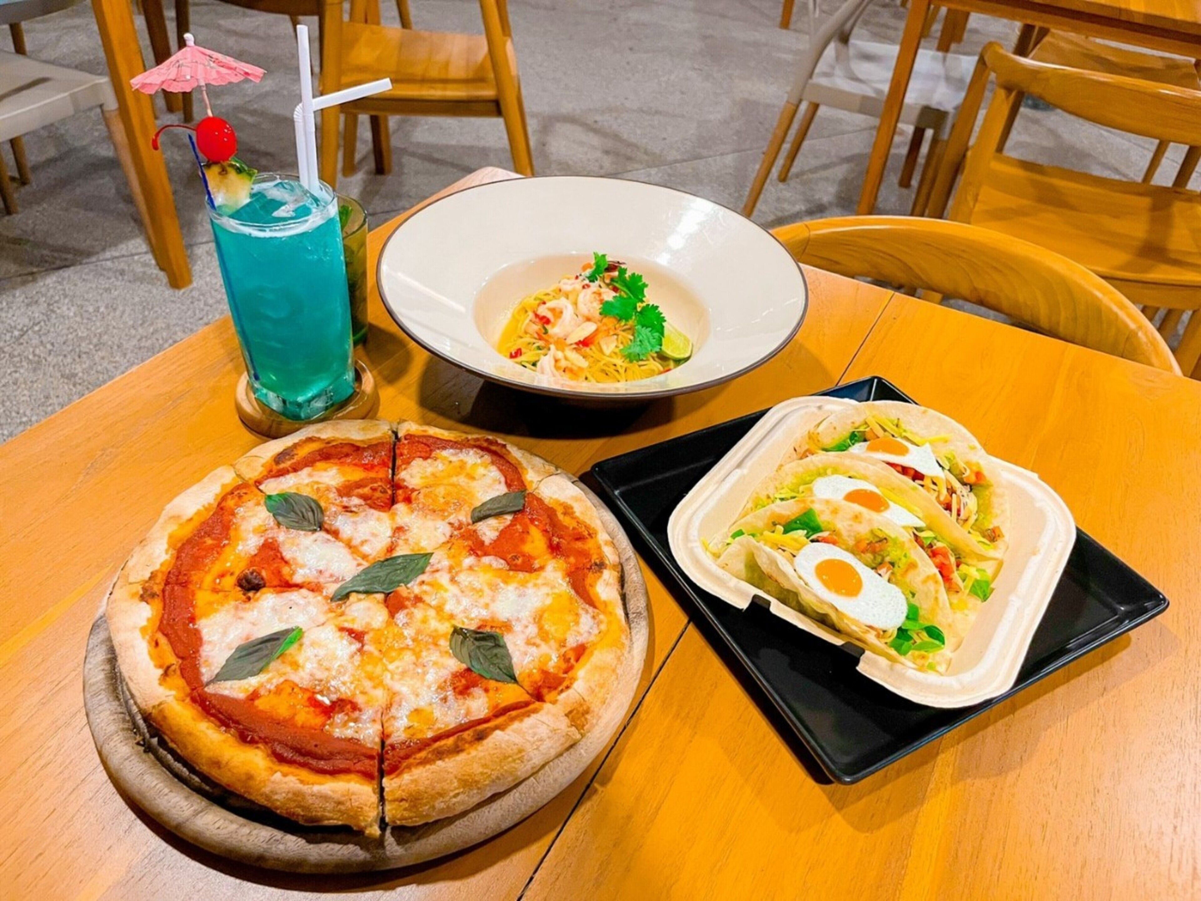 A pizza, a bowl of soup, and a container of tacos on a wooden table.