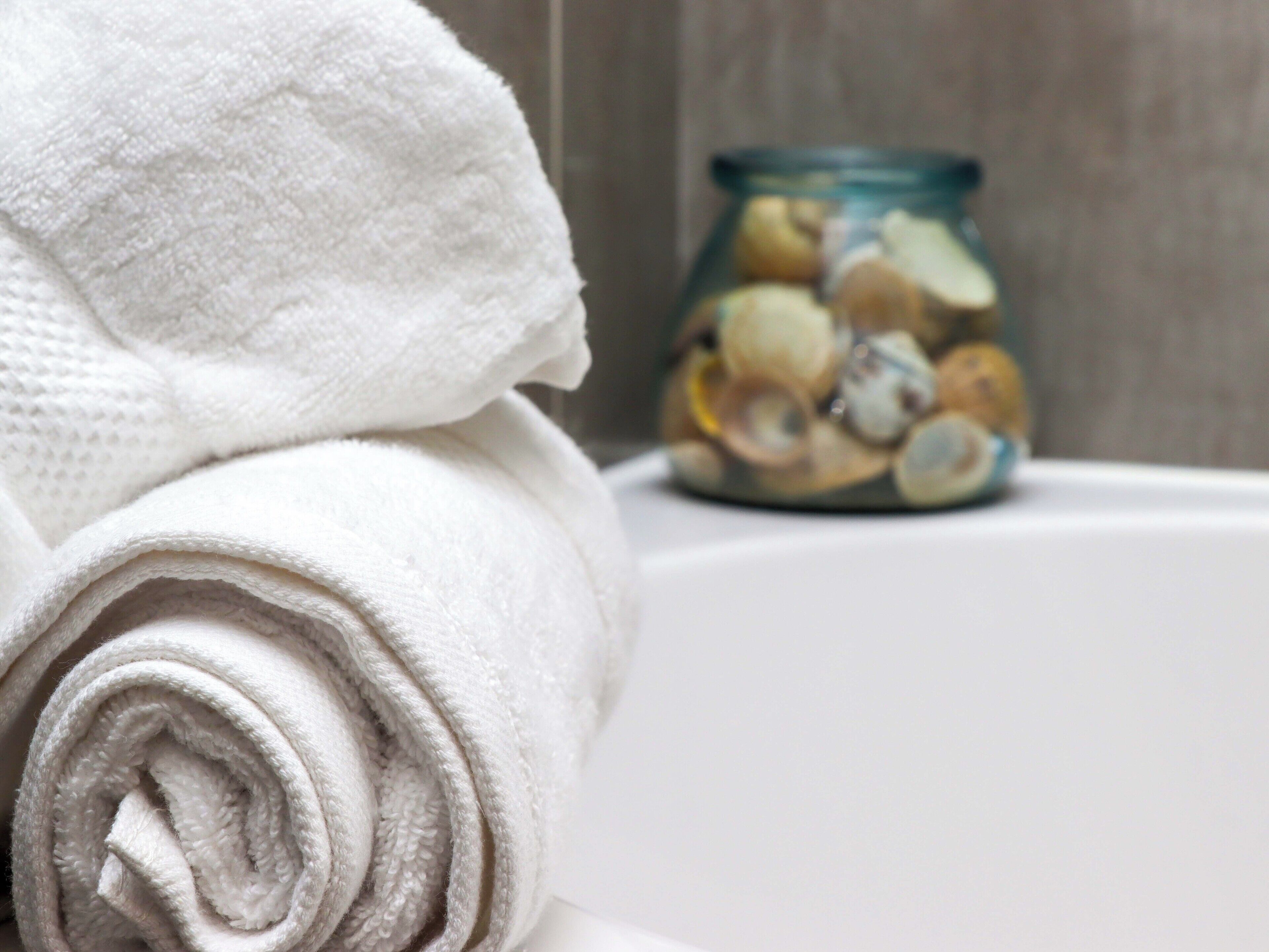 A rolled white towel on a white surface with a glass jar containing various items in the background.