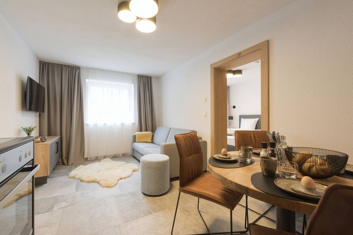 Ein modernes Wohnzimmer mit einer Couch, einem Esstisch mit Stühlen, einem am Wand befestigten Flachbildfernseher und einem großen Spiegel, der den Raum widerspiegelt.