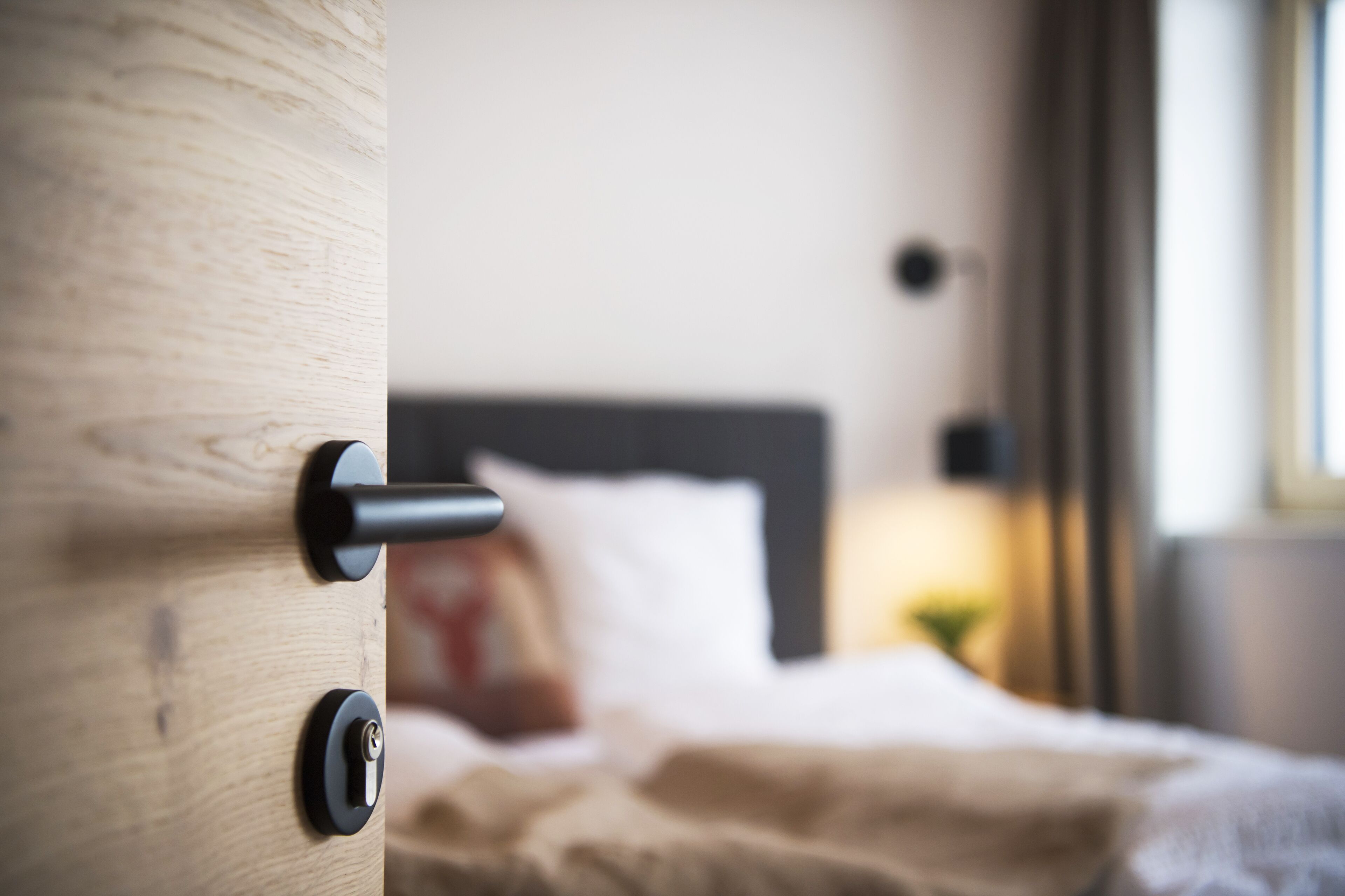 A close-up of a wooden door with a keyhole and a handle, leading to a blurred bedroom interior with a bed and a bedside lamp.