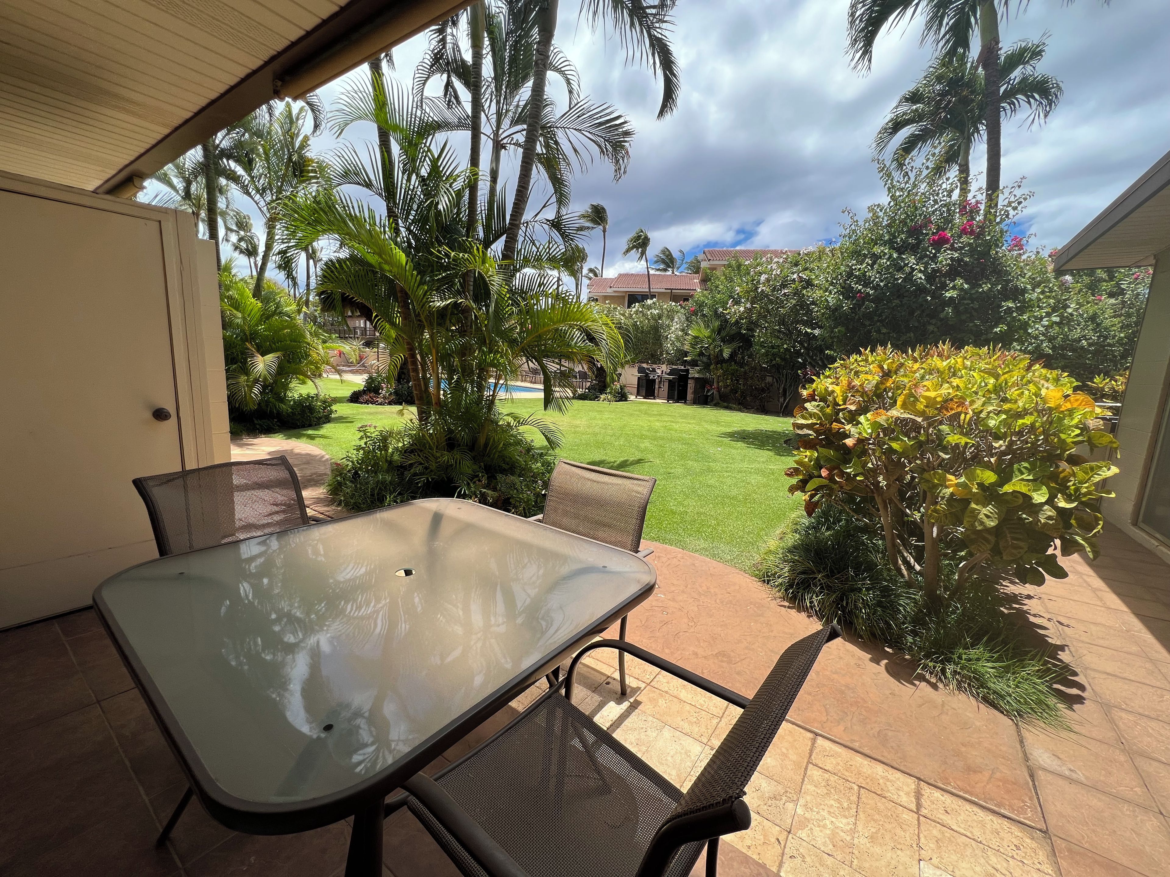 An outdoor patio with a glass table and chairs, surrounded by palm trees and a well-maintained lawn.