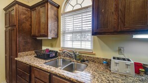 Una cocina con armarios de madera, encimera de granito, pileta doble, ventana con persianas y un tostador.