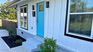 Una casa con puerta azul, revestimiento blanco y un pequeño jardín con arbustos.