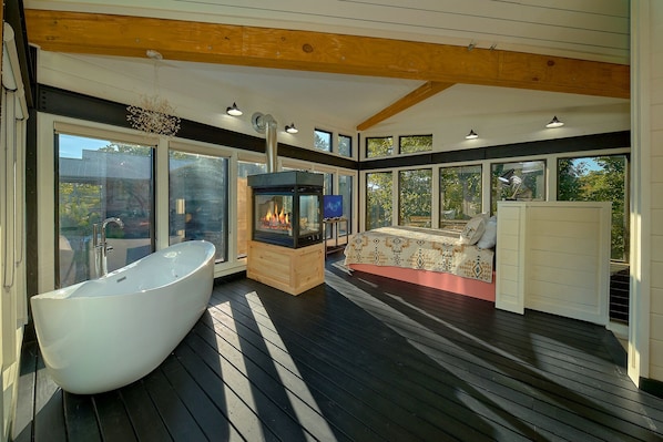 A modern bathroom with a freestanding bathtub, a wood-burning stove, and a large window offering a view of the outdoors.