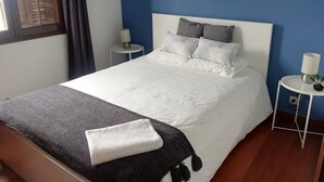 A neatly made bed with white linens, a grey throw blanket, and a folded white towel. There are two bedside tables with lamps, one on each side of the bed. The room has blue walls and a window with blinds. - Casa da Travessa (Horta)