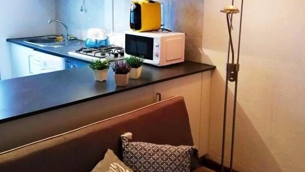 A compact living space with a kitchenette, a sofa with a 'HAPPY PLACE' pillow, and a floor lamp. - Casa da Travessa (Horta)